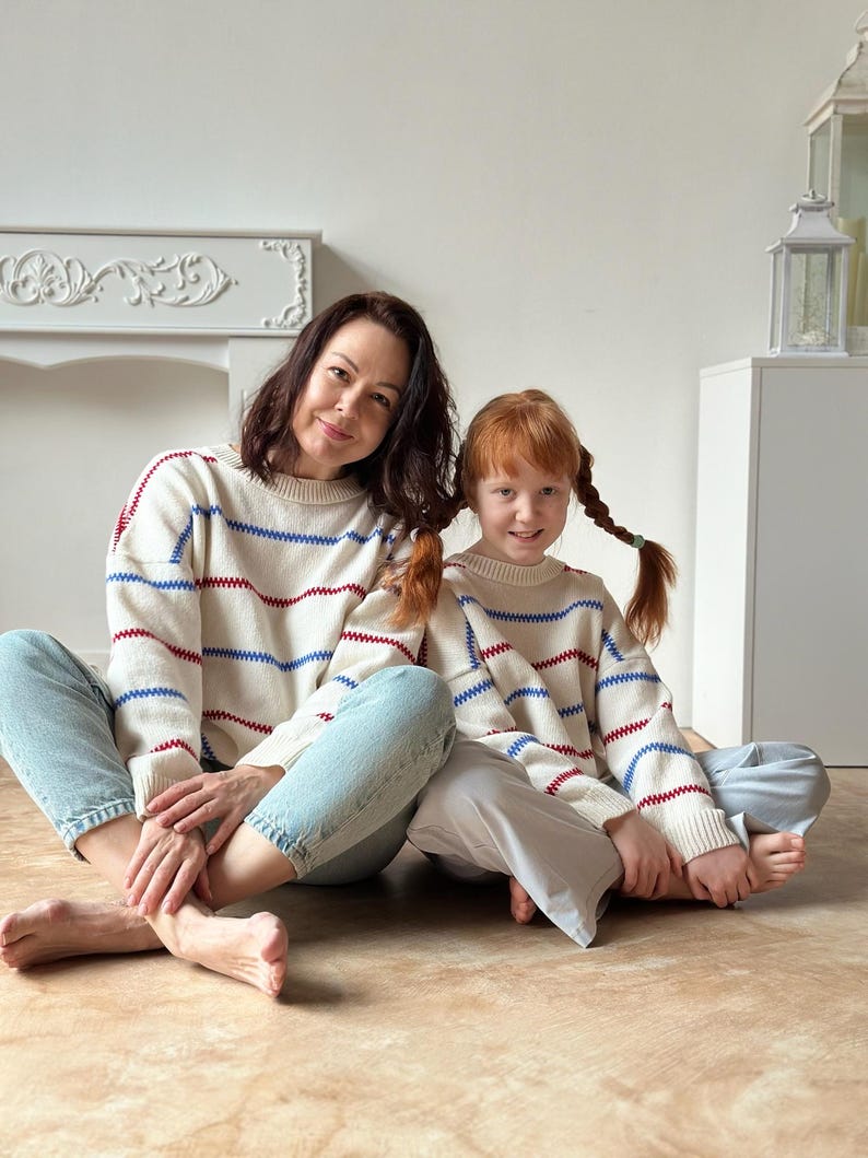 May include: Two people wearing matching cream-colored sweaters with red and blue horizontal stripes. They are sitting on a light brown surface. The sweaters have a crew neck and long sleeves. The person on the left is wearing blue jeans, and the person on the right is wearing light blue pants.