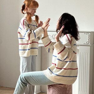 May include: Two people wearing matching cream-colored sweaters with red and blue stripes. The sweaters have a ribbed hem and cuffs. They are wearing light blue jeans. One person is sitting on a pink velvet stool.