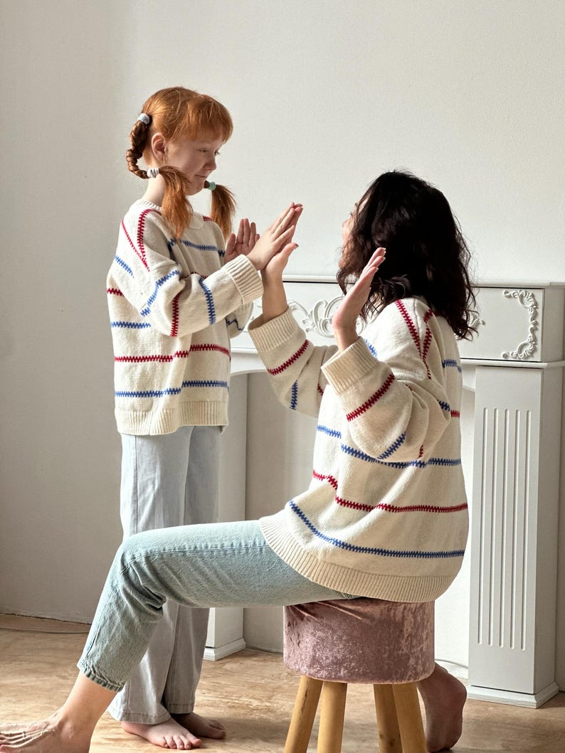 May include: Two people wearing matching cream-colored sweaters with red and blue stripes. The sweaters have a ribbed hem and cuffs. They are wearing light blue jeans. One person is sitting on a pink velvet stool.