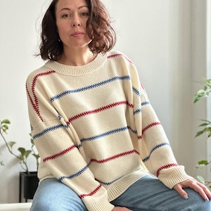 May include: Cream-colored sweater with horizontal blue and red stripes. The sweater has a crew neck and long sleeves. The stripes are evenly spaced across the body and sleeves. The sweater is paired with blue jeans.