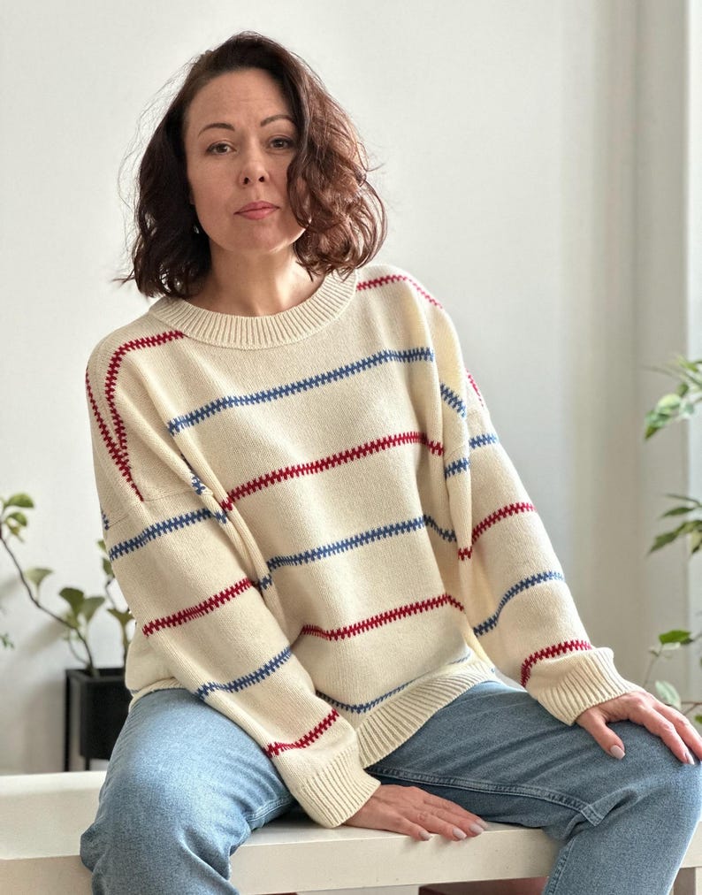 May include: Cream-colored sweater with horizontal blue and red stripes. The sweater has a crew neck and long sleeves. The stripes are evenly spaced across the body and sleeves. The sweater is paired with blue jeans.