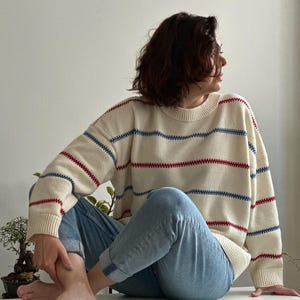 May include: Cream-colored sweater with horizontal red and blue stripes. The sweater has a crew neck and long sleeves. The person is wearing light blue jeans.