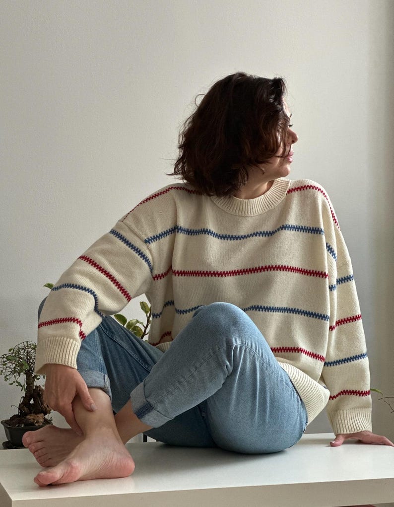 May include: Cream-colored sweater with horizontal red and blue stripes. The sweater has a crew neck and long sleeves. The person is wearing light blue jeans.