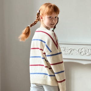 May include: Cream-colored sweater with horizontal red and blue stripes. The sweater has a classic crew neck and long sleeves. The stripes are evenly spaced across the body and sleeves. The sweater is worn by a person with red hair in braids.