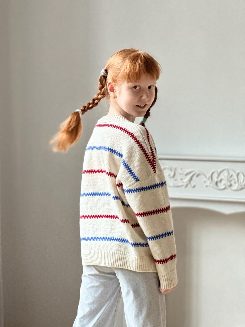 May include: Cream-colored sweater with horizontal red and blue stripes. The sweater has a classic crew neck and long sleeves. The stripes are evenly spaced across the body and sleeves. The sweater is worn by a person with red hair in braids.
