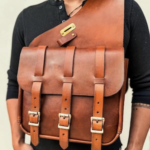 Op de afbeelding: Een bruine leren schoudertas met drie voorste riemen en messing gespen. De tas heeft een klepsluiting en een schouderriem. De tas wordt gedragen door een persoon die een zwart shirt met lange mouwen draagt.
