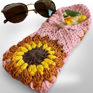 May include: A pink crocheted sunglasses case with a sunflower design. The case has a brown button closure and a floral lining.