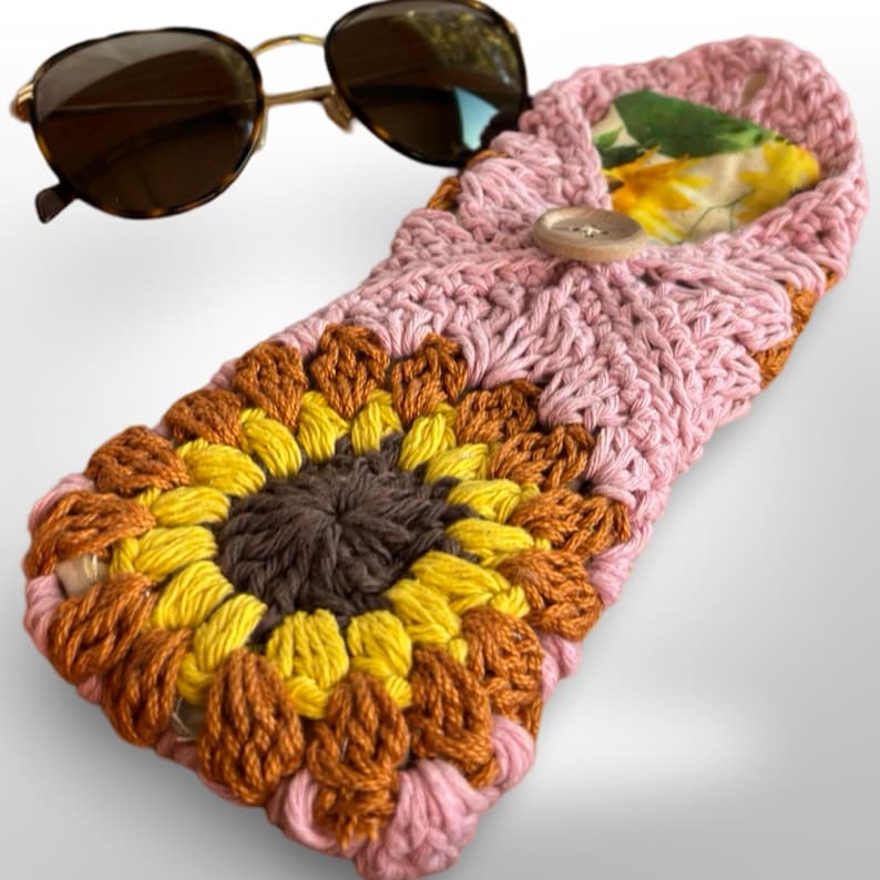 May include: A pink crocheted sunglasses case with a sunflower design. The case has a brown button closure and a floral lining.