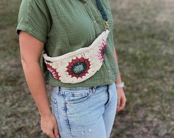 May include: A woman wearing a green shirt and blue jeans is wearing a crocheted fanny pack with pink, red, and green accents. The fanny pack is worn across the body and has a long strap.