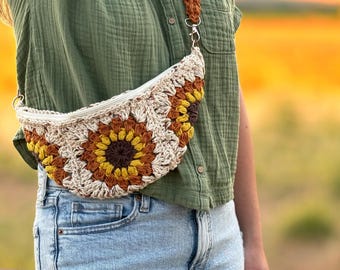 Könnte beinhalten: Eine gehäkelte Sonnenblumen-Bauchtasche mit weißem Hintergrund und braunen, gelben und orangefarbenen Akzenten. Die Tasche wird um die Taille getragen und hat einen langen Riemen.