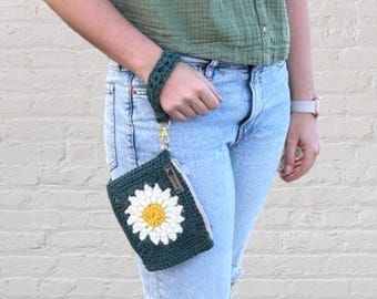 May include: A green crocheted purse with a white daisy flower design. The purse has a gold chain strap and is being held by a person wearing blue jeans.