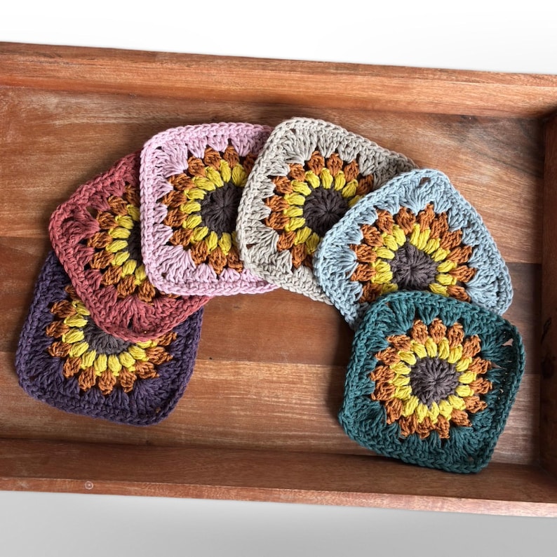 May include: A set of six crocheted sunflower squares in various colors, including purple, pink, beige, blue, green, and brown. The squares are arranged in a rainbow pattern and are displayed in a wooden tray.
