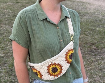 May include: A crocheted sunflower-patterned crossbody bag with a white strap. The bag is worn by a person wearing a green shirt.