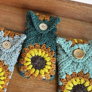 May include: Four crocheted phone cases with sunflower designs. The cases are in shades of blue, green, and brown. Each case has a wooden button closure.