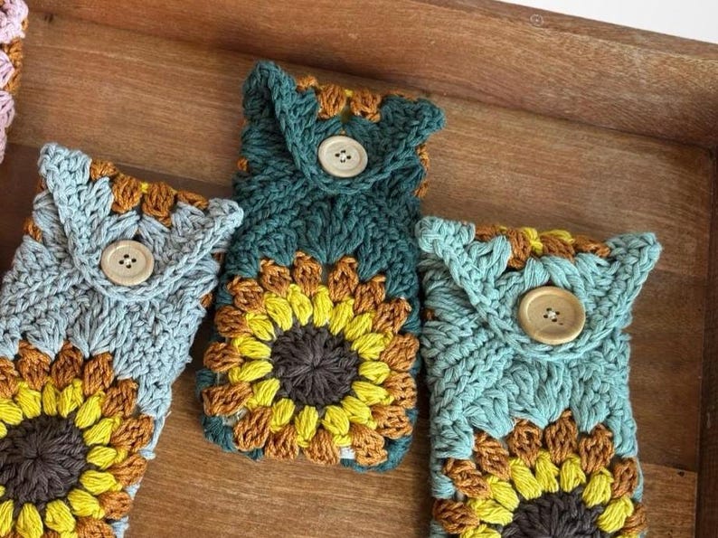May include: Four crocheted phone cases with sunflower designs. The cases are in shades of blue, green, and brown. Each case has a wooden button closure.