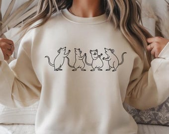 May include: Cream-colored sweatshirt featuring a black line drawing of five mice holding hands. The mice are in various poses, with some standing upright. The sweatshirt has long sleeves and a crew neck.
