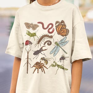 May include: A cream-colored t-shirt featuring a detailed graphic print of various insects and invertebrates, including a moth, earthworm, monarch butterfly, grasshopper, centipede, ladybug, dragonfly, beetle, spider, wasp, stick insect, and mantis.