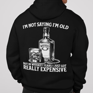 Può includere: Felpa con cappuccio nera con grafica bianca. La grafica presenta una bottiglia di whisky e un bicchiere di whisky, con la scritta "I'M NOT SAYING I'M OLD BUT IN WHISKEY YEARS I JUST GOT REALLY EXPENSIVE."