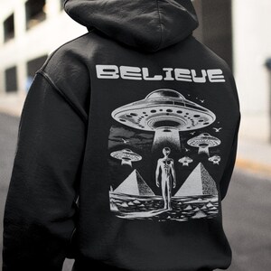 Pode incluir: Camisola de moletom com capuz preta com uma estampa gr&aacute;fica de um OVNI, um alien&iacute;gena e pir&acirc;mides. O texto "BELIEVE" est&aacute; impresso na parte de tr&aacute;s da camisola.