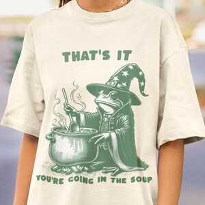 Puede incluir: Camiseta color crema con una ilustración verde de una rana vestida de mago, removiendo una olla de sopa. El texto "THAT'S IT" y "YOU'RE GOING IN THE SOUP" están impresos encima y debajo del gráfico de la rana.