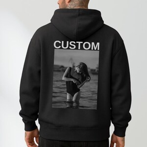 May include: Black hoodie with the word "CUSTOM" printed in white across the upper back. A black and white photo of a person in a swimsuit in the water is printed below the word "CUSTOM". The hoodie has a hood and a relaxed fit.