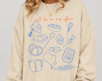 May include: A beige crewneck sweatshirt featuring the text I will be in my office in orange lettering. The front displays blue line art illustrations of baking tools including a mixer, whisk, measuring cup, rolling pin, and a jar labeled flour.