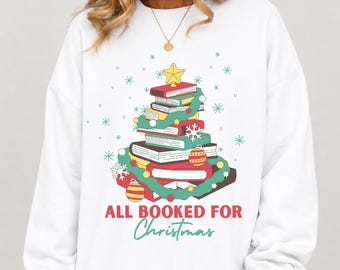 May include: A white long-sleeve sweatshirt featuring a graphic of a Christmas tree made of stacked books, decorated with a yellow star, green garland, and ornaments. The text reads ALL BOOKED FOR Christmas in red and green lettering.