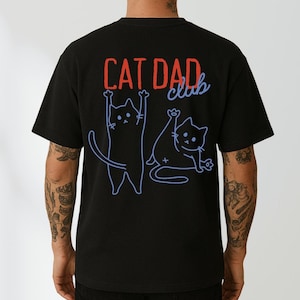 Puede incluir: Camiseta negra con las palabras "CAT DAD club" en rojo y azul. Dos ilustraciones de gatos azules aparecen debajo del texto. Un gato está de pie con las patas levantadas y el otro está sentado con una pata levantada.