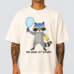 Könnte beinhalten: Ein cremefarbenes Kurzarm-T-Shirt mit einer Grafik eines Waschbären, der ein blaues Visier trägt und einen blauen Pickleball-Schläger sowie einen gelben Pickleball hält. Der Text unter dem Waschbären lautet WE DINK AT DAWN in schwarzen Buchstaben.