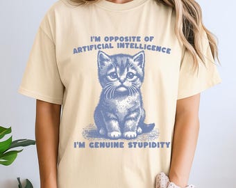 Pode incluir: Uma t-shirt de manga curta na cor creme com um gráfico azul de um gatinho. O texto acima do gatinho diz I'M OPPOSITE OF ARTIFICIAL INTELLIGENCE e o texto abaixo do gatinho diz I'M GENUINE STUPIDITY em uma fonte azul.