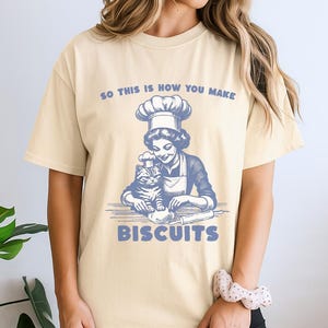 Puede incluir: Camiseta color crema con una ilustración azul de un chef con un gato, amasando masa. El texto superior dice "SO THIS IS HOW YOU MAKE" y debajo "BISCUITS". La camiseta tiene mangas cortas y un corte holgado.