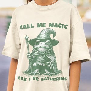 May include: Cream-colored t-shirt with a green graphic of a frog dressed as a wizard, holding a staff. The text "CALL ME MAGIC" is above the frog, and "CUZ I BE GATHERING" is below. The frog wears a pointed hat and cape.