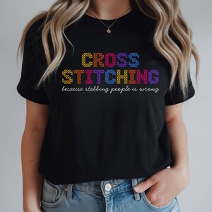 May include: Black t-shirt featuring the words "CROSS STITCHING" in a colourful cross-stitch design. The phrase "because stabbing people is wrong" is printed below in white.