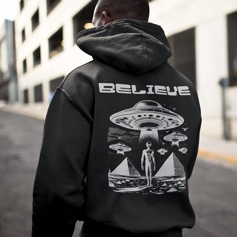 Pode incluir: Camisola de moletom com capuz preta com uma estampa gr&aacute;fica de um OVNI, um alien&iacute;gena e pir&acirc;mides. O texto "BELIEVE" est&aacute; impresso na parte de tr&aacute;s da camisola.