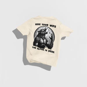 May include: Cream-colored t-shirt featuring a black and white illustration of a werewolf howling at the moon. The text "HIDE YOUR WIFE" and "THE ALPHA IS HERE" are printed above and below the graphic, respectively.