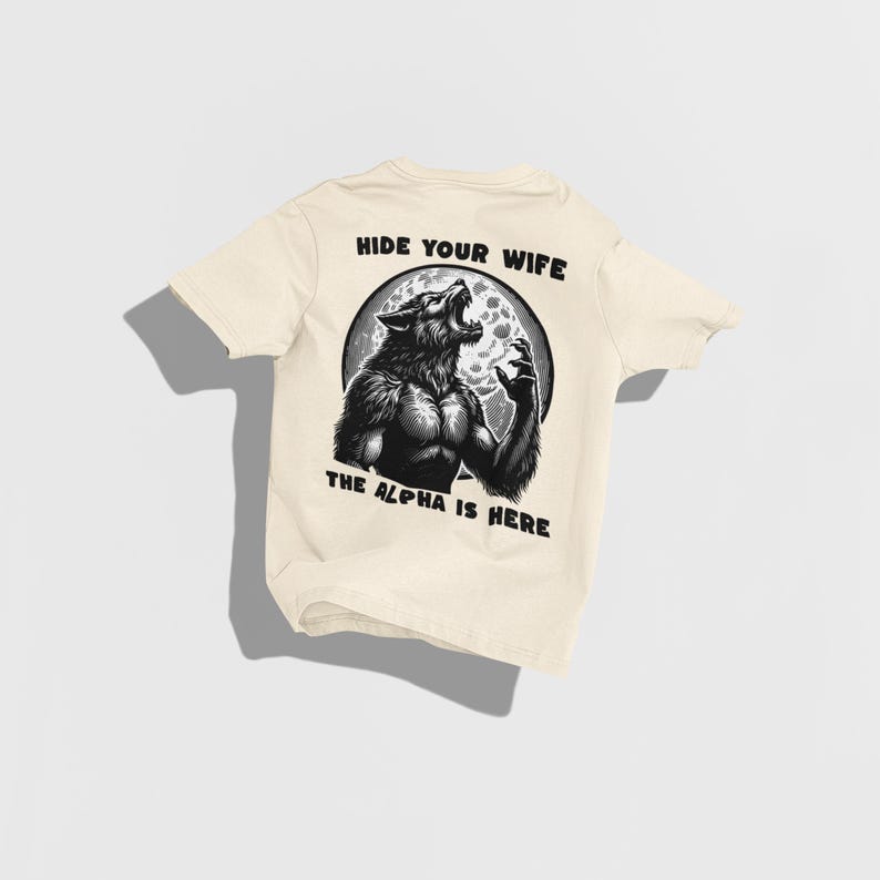 May include: Cream-colored t-shirt featuring a black and white illustration of a werewolf howling at the moon. The text "HIDE YOUR WIFE" and "THE ALPHA IS HERE" are printed above and below the graphic, respectively.