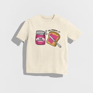 May include: Cream-colored t-shirt featuring a graphic of a jar labeled "THE GOSPEL" and a slice of toast with pink spread and a knife. The text "GO SPREAD IT" is above the toast.