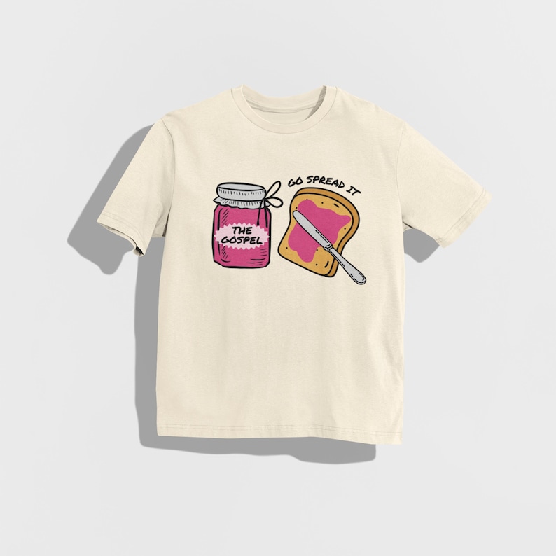 May include: Cream-colored t-shirt featuring a graphic of a jar labeled "THE GOSPEL" and a slice of toast with pink spread and a knife. The text "GO SPREAD IT" is above the toast.