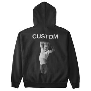 May include: Black hoodie with a hood and long sleeves. The word "CUSTOM" is printed in white across the upper back. A black and white image of a person making a peace sign is below the word "CUSTOM".