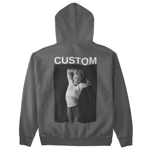 May include: A charcoal gray hoodie with a hood and a front pocket. The back features a black and white image of a person making a peace sign, with the word "CUSTOM" printed above the image.