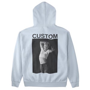 May include: A light blue hoodie with a black and white image of a person making a peace sign. The word "CUSTOM" is printed in black above the image. The hoodie has a hood and long sleeves.