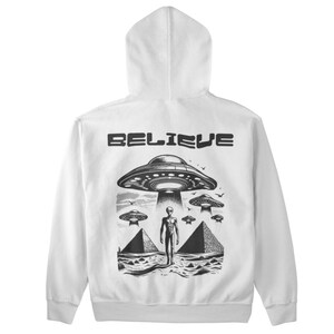 Pode incluir: Camisola de capuz branca com uma estampa gr&aacute;fica em preto e branco de um OVNI e uma figura alien&iacute;gena. O texto "BELIEVE" est&aacute; impresso acima do gr&aacute;fico.