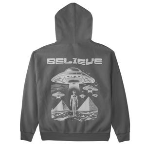 Pode incluir: Uma camisola de capuz cinzenta antracite com uma estampa gr&aacute;fica branca de um OVNI, pir&acirc;mides e uma figura alien&iacute;gena. O texto "BELIEVE" est&aacute; impresso acima do gr&aacute;fico.