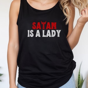 Op de afbeelding: Zwart mouwloos shirt met de tekst "SATAN IS A LADY" in rood en wit. Het woord "SATAN" is rood met een duivelshoornontwerp erboven. De tekst staat gecentreerd op de voorkant van het mouwloze shirt.