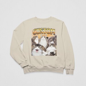 May include: A beige crewneck sweatshirt featuring a graphic print of five fluffy cats. Above the cats, the word CUSTOM is printed in a bold, yellow-outlined font, with the words Your text printed in smaller white letters directly underneath.