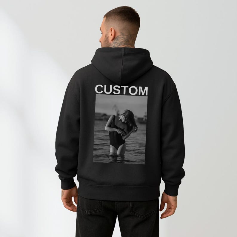 May include: Black hoodie with the word "CUSTOM" printed in white across the upper back. A black and white photo of a person in a swimsuit in the water is printed below the word "CUSTOM". The hoodie has a hood and a relaxed fit.