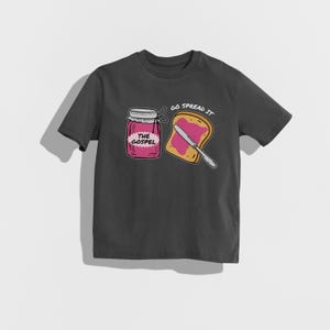 May include: Dark gray t-shirt featuring a graphic of a jar of pink jam labeled "THE GOSPEL" next to a slice of toast with jam and a knife. The text "GO SPREAD IT" is above the toast.