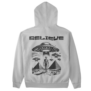 Pode incluir: Uma jaqueta de moletom cinza com capuz com uma estampa gr&aacute;fica preta de um alien&iacute;gena em p&eacute; na frente de uma pir&acirc;mide com um OVNI acima. O texto "BELIEVE" &eacute; impresso acima do gr&aacute;fico.