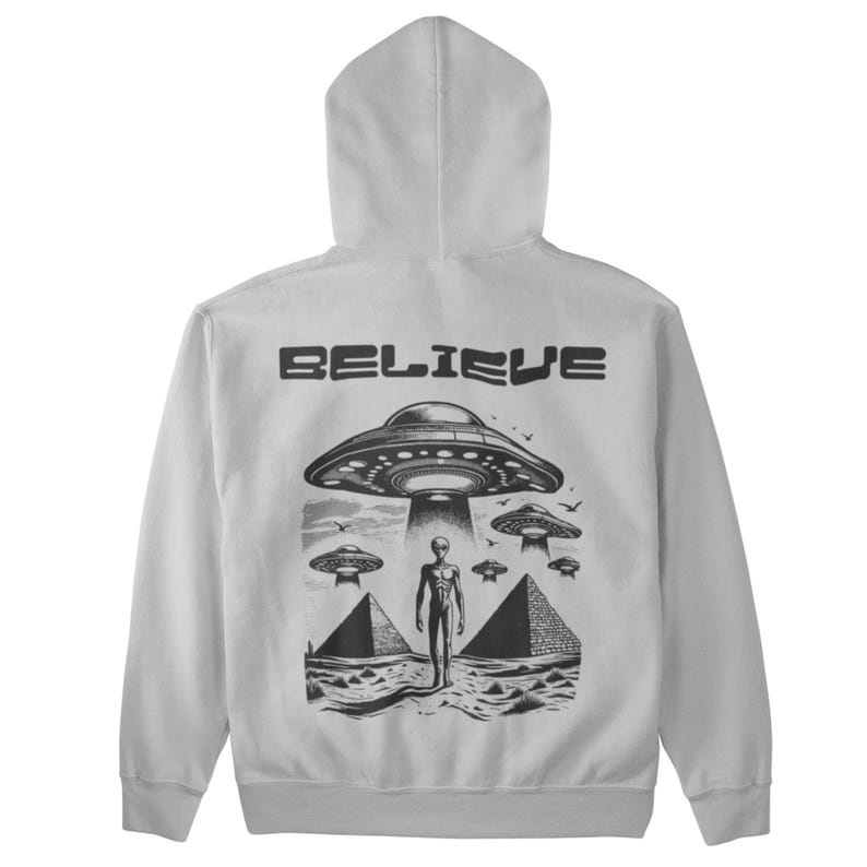 Pode incluir: Uma jaqueta de moletom cinza com capuz com uma estampa gr&aacute;fica preta de um alien&iacute;gena em p&eacute; na frente de uma pir&acirc;mide com um OVNI acima. O texto "BELIEVE" &eacute; impresso acima do gr&aacute;fico.