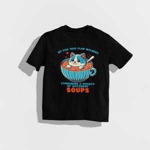 May include: Black t-shirt featuring a white cartoon cat in a bowl of soup. The text on the shirt reads "My five year plan includes consuming a variety of different soups."